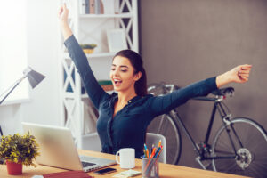 Winning every day. Attractive young woman stretching out hands and looking excited while sitting at her working place and looking at laptop